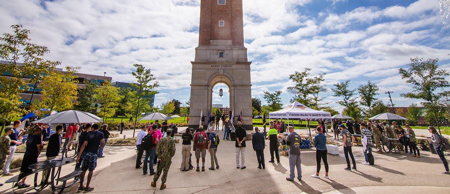 Large group of people some in military uniforms gather in front of Elliott Tower on Oakland University's campus